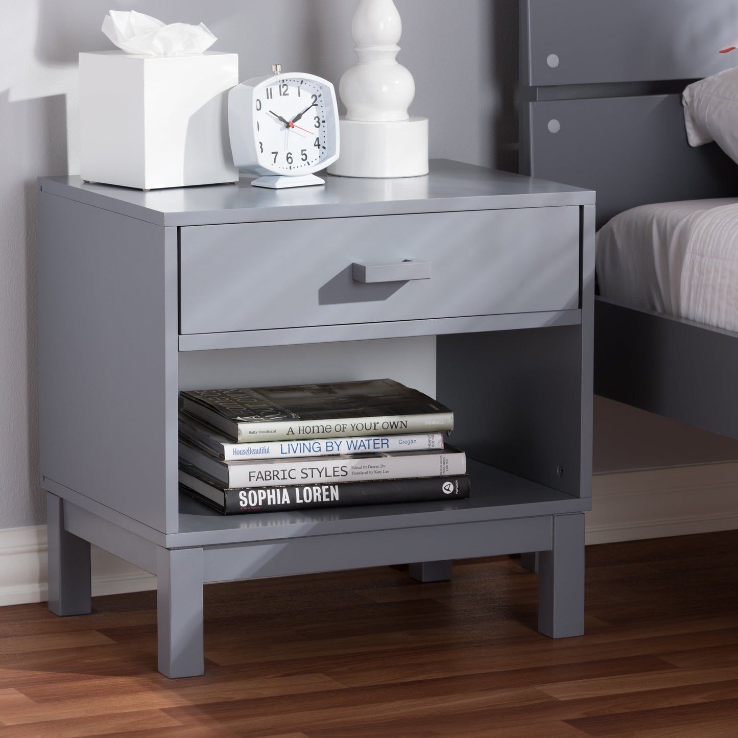 Grey bedside table featuring a drawer and open shelf holding stacked books and decor.