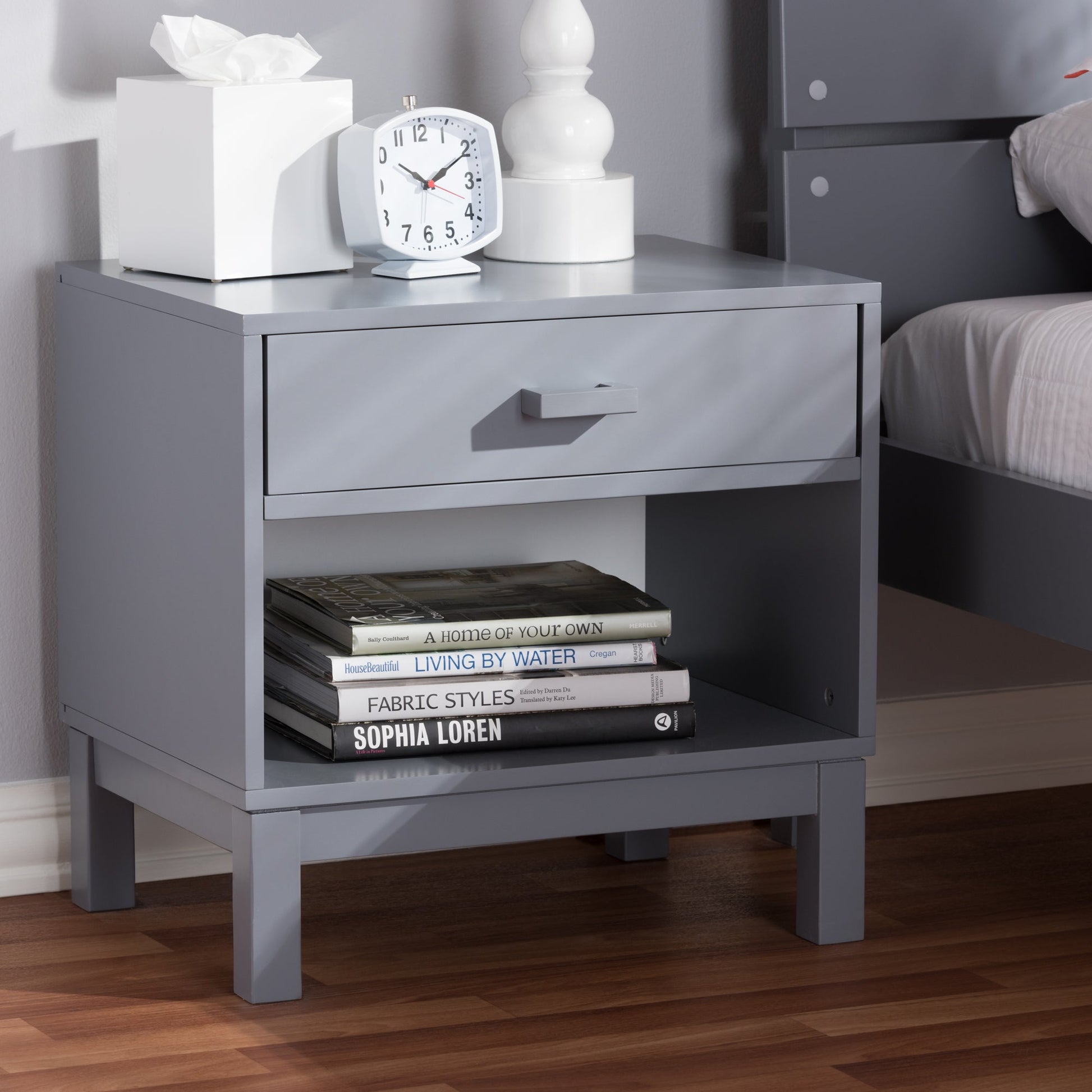 Grey bedside table featuring a drawer and open shelf holding stacked books and decor.